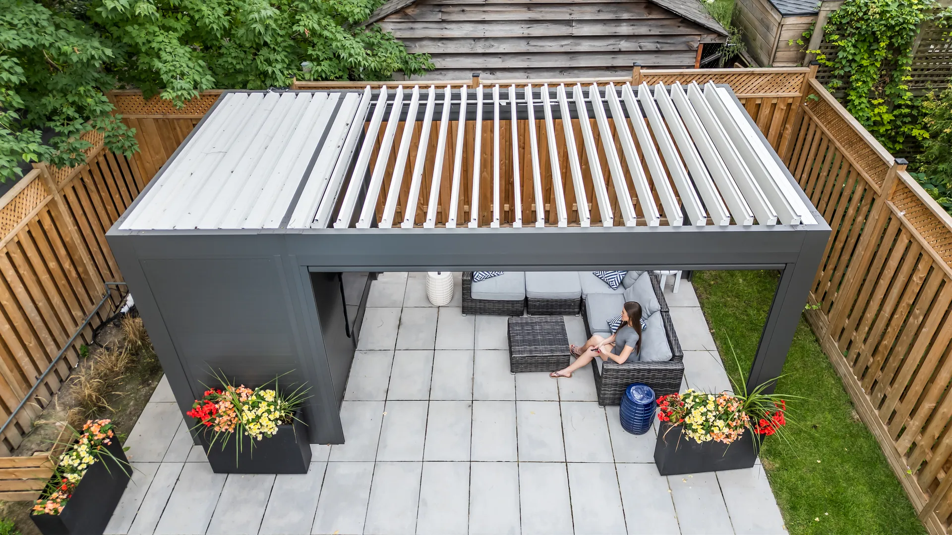 Aerial view of a grey aluminum pergola with white louvers sized to fit a fenced backyard patio with a lounge sofa