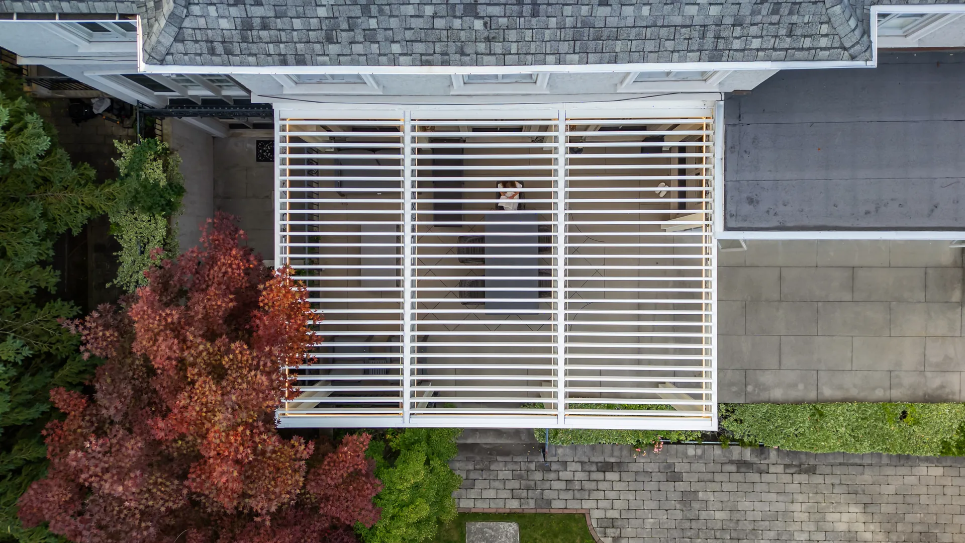 Top-down view of a white attached pergola with three louvered roof zones over a dining table beside the house