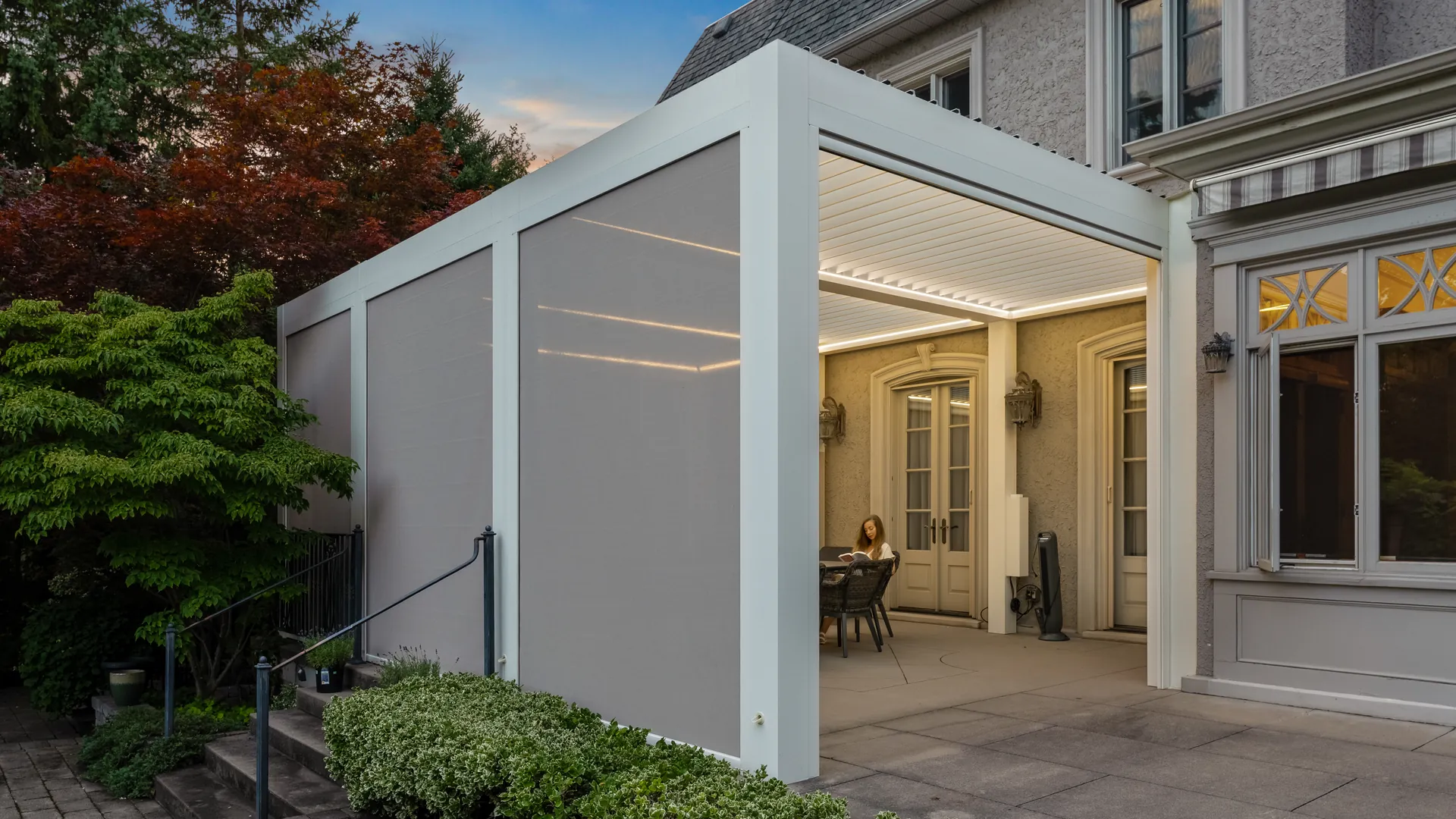 White attached louvered pergola with three beige pergola screens lowered along one side and LED lights over a dining table
