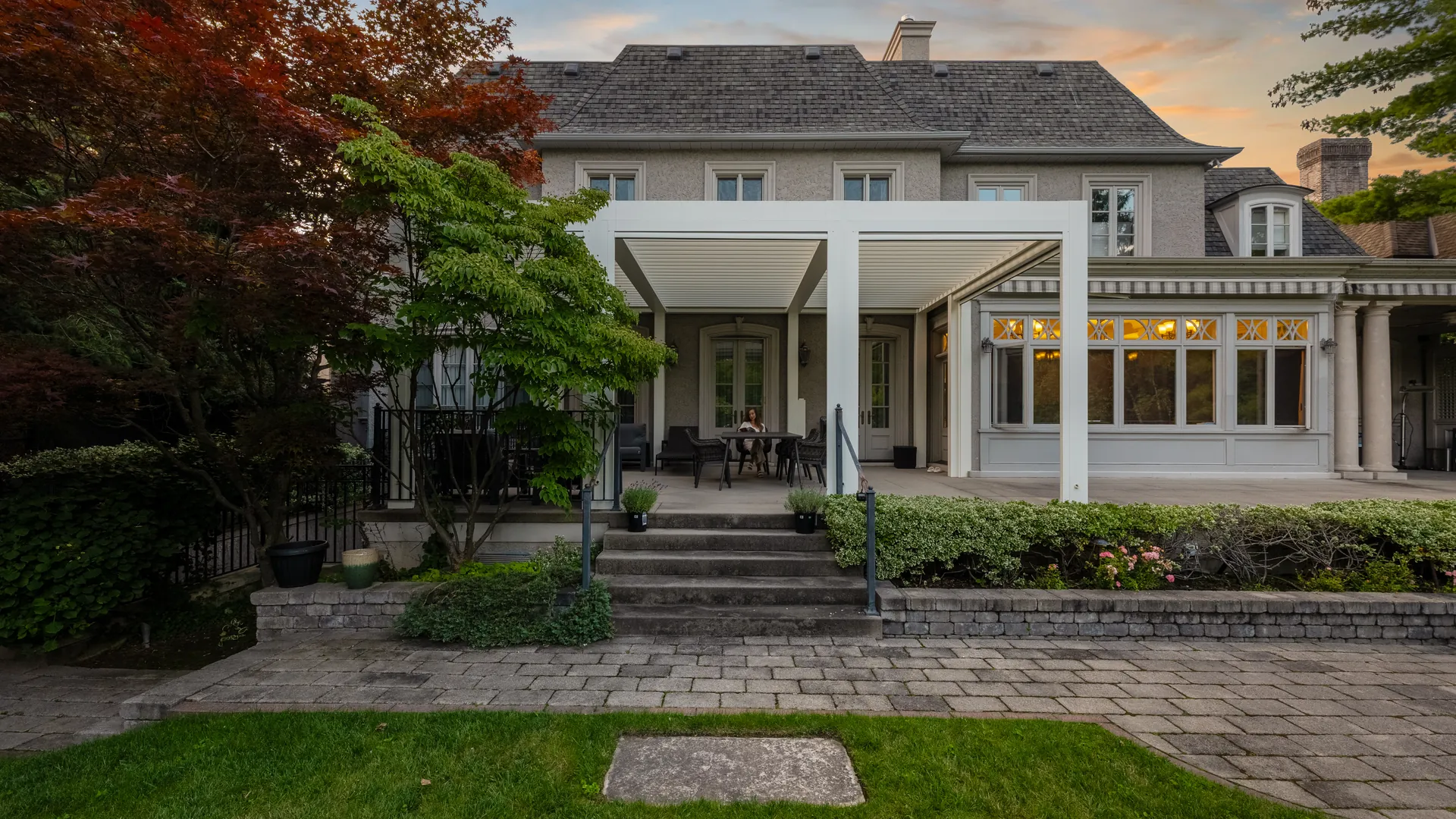 White attached aluminum louvered pergola on the back of a stone house, over a raised patio with steps to the garden