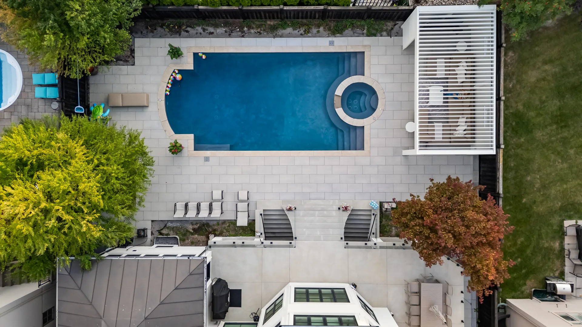 Aerial view of a fenced backyard with a pool, round spa and a white louvered pergola in the corner, surrounded by trees