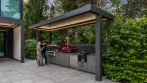Covered BBQ area seen from the side: aluminum pergola with an LED-lit roof over a built-in grill, a pizza oven and grey cabinets beside a hedge
