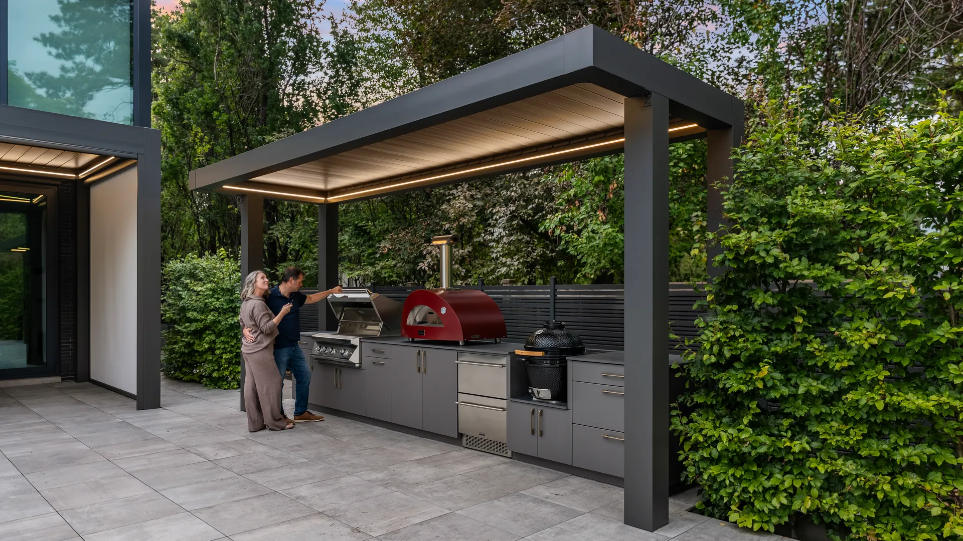 Covered BBQ area seen from the side: aluminum pergola with an LED-lit roof over a built-in grill, a pizza oven and grey cabinets beside a hedge