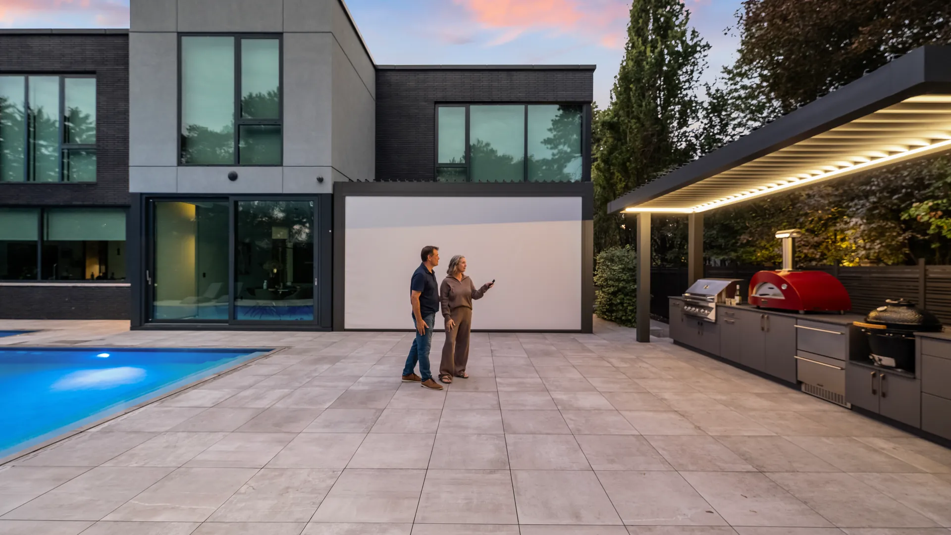Covered outdoor kitchen design next to a modern home and pool at dusk, with a white zip screen lowered on the house pergola