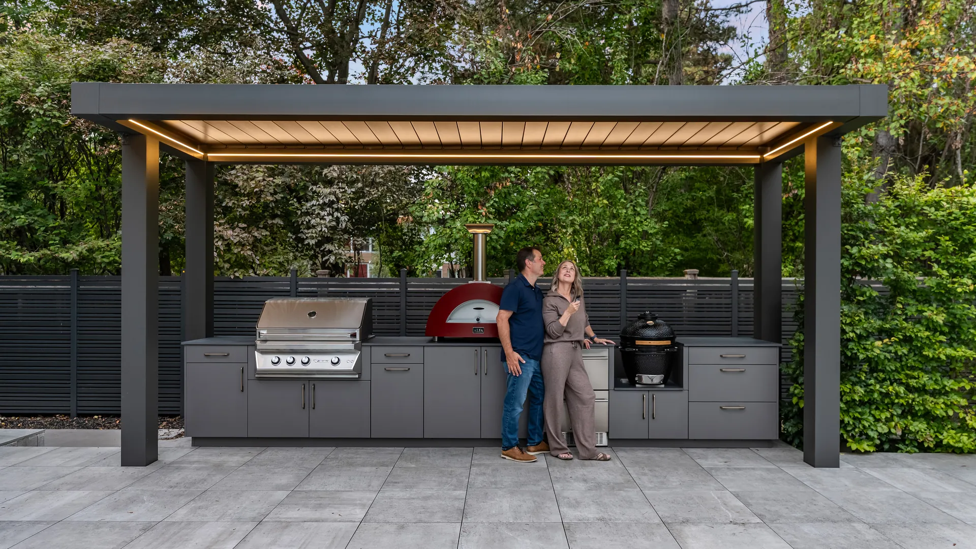 Covered outdoor kitchen idea: a freestanding Umbra Grey pergola with white louvers and LED lights over a grill, red pizza oven and kamado, with a couple at the counter
