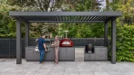 Outdoor kitchen under a louvered pergola with the louvers open to the sky and a couple cooking at the built-in grill
