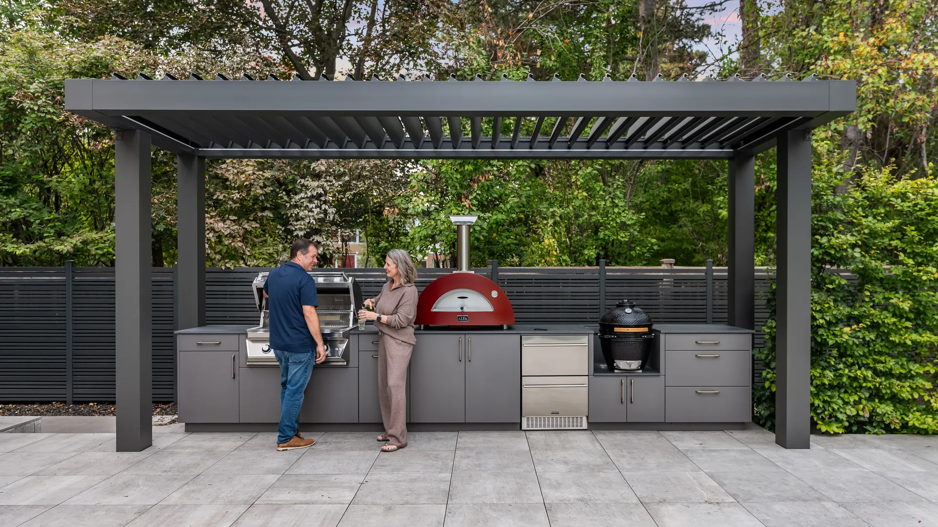 Outdoor kitchen under a louvered pergola with the louvers open to the sky and a couple cooking at the built-in grill
