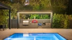 Covered outdoor kitchen at night under a grey aluminum pergola with white louvers and warm LED lights, a red pizza oven and a lit pool in front, Thornhill, Ontario
