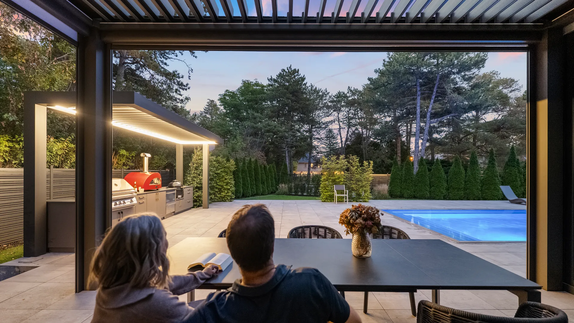 Couple at the dining table under the house pergola, looking out at the lit outdoor kitchen pergola and the pool at dusk