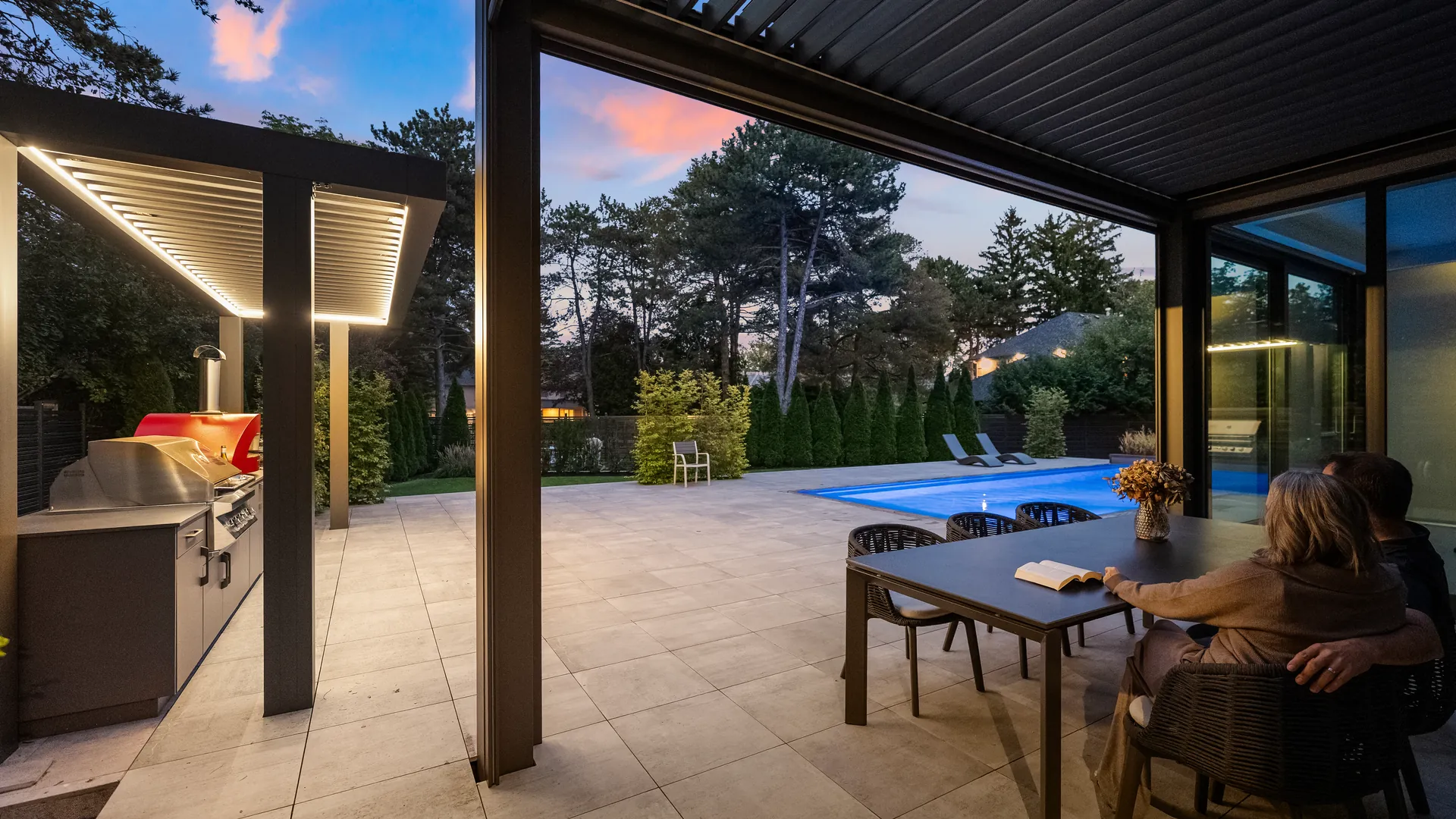 Couple dining under a dark aluminum louvered pergola at dusk, next to an LED-lit pergola over an outdoor kitchen and a pool