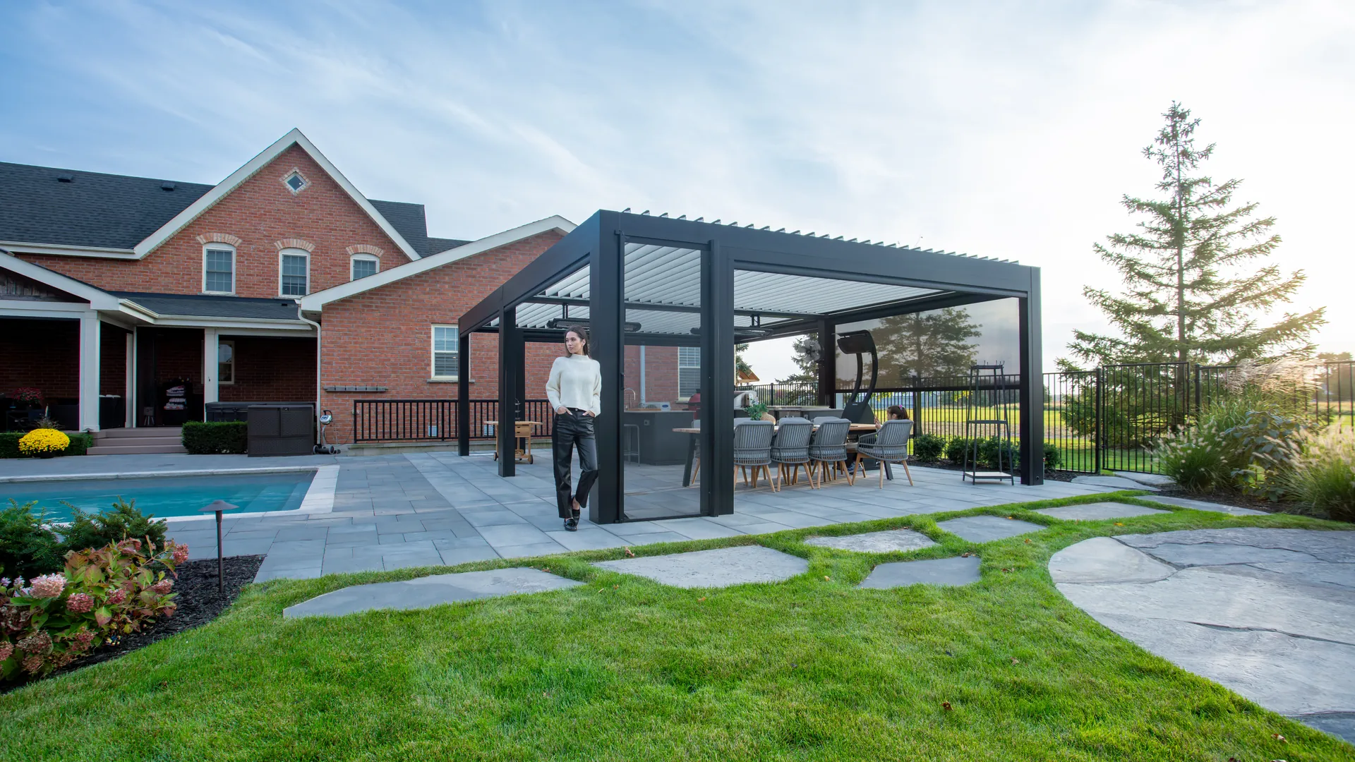 Freestanding black aluminum louvered pergola with an outdoor dining area on a stone patio beside a pool and brick house