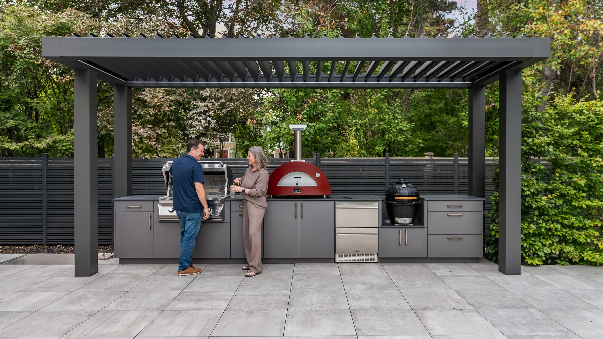 Homeowners at an outdoor kitchen under a freestanding dark aluminum louvered pergola on a large-format paver patio