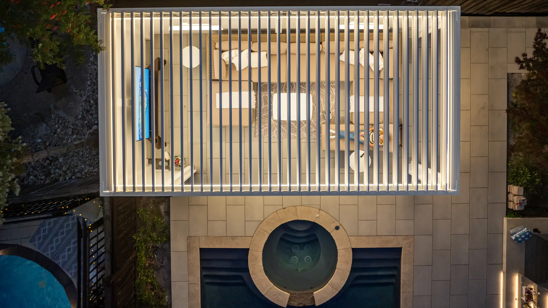 Top-down night view of a white louvered pergola with LED lighting over a lounge next to a hot tub and pool
