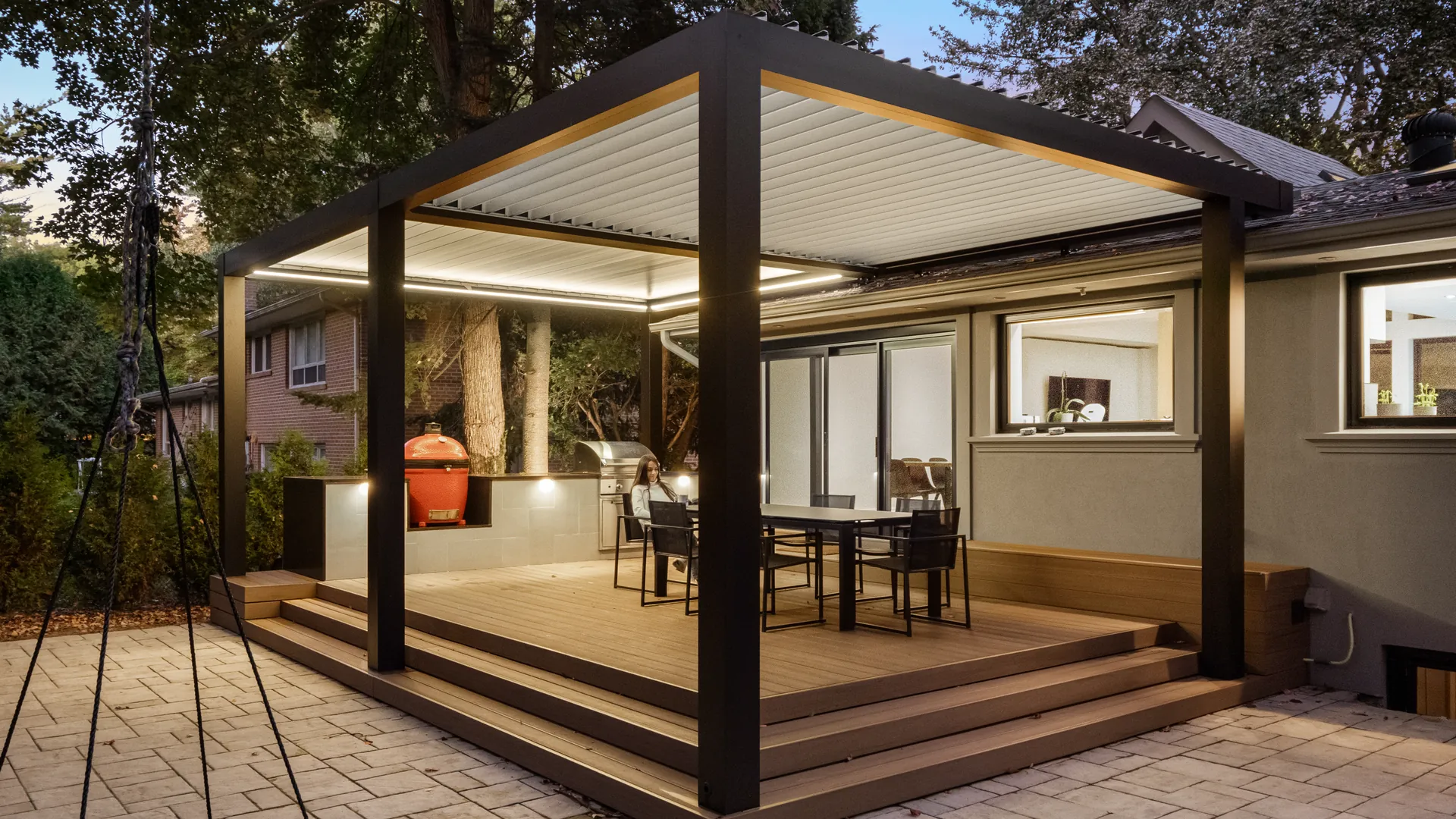 Black aluminum pergola with white louvers and LED lighting over a raised composite deck with a dining set at dusk
