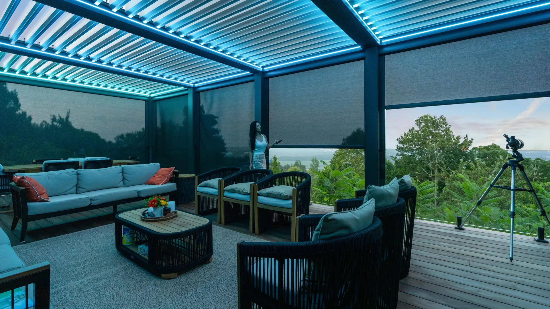 Woman using a remote to set ZIP screens on a louvered pergola with blue LED lighting over a lounge on a deck