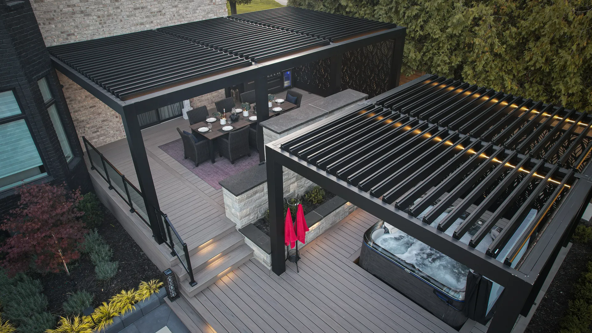 Aerial view of a modern backyard pergola design: two black louvered pergolas on a two-tier deck, dining area above and hot tub below