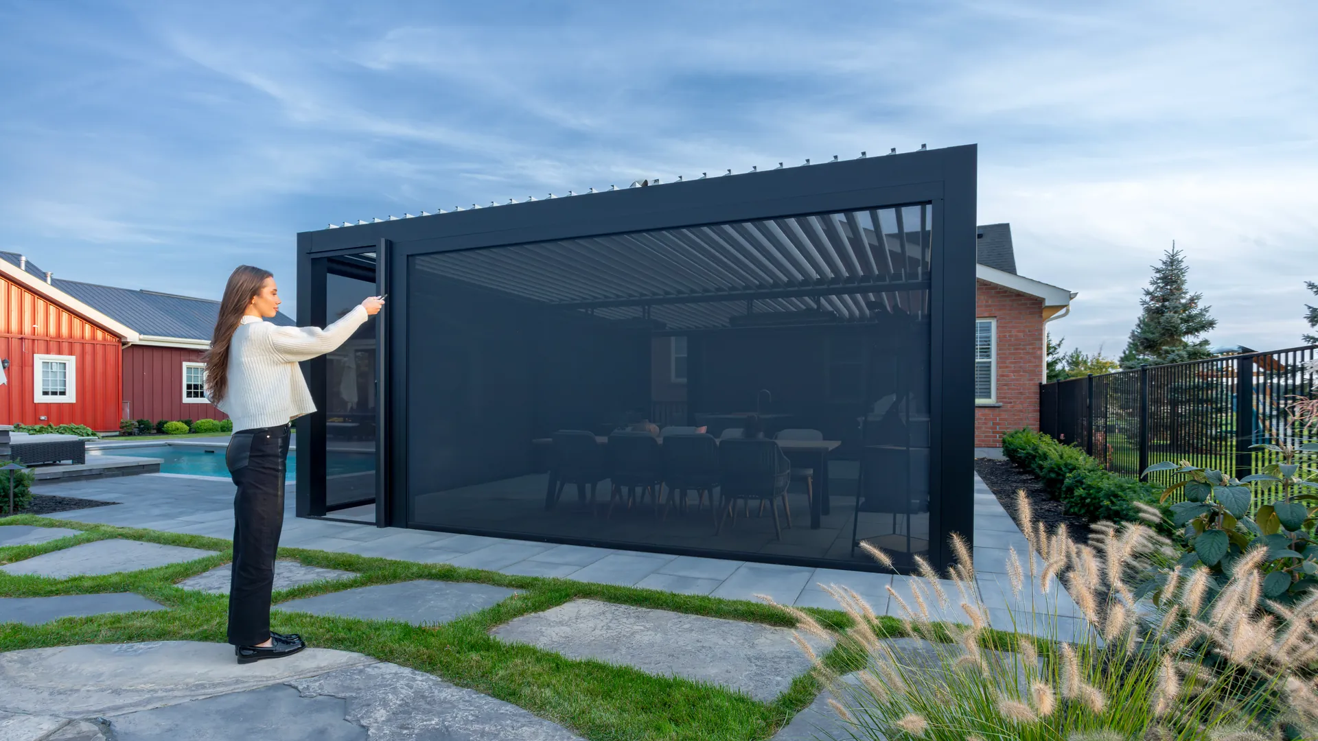 Woman lowering motorized pergola screens with a handheld remote on a black louvered pergola beside a pool