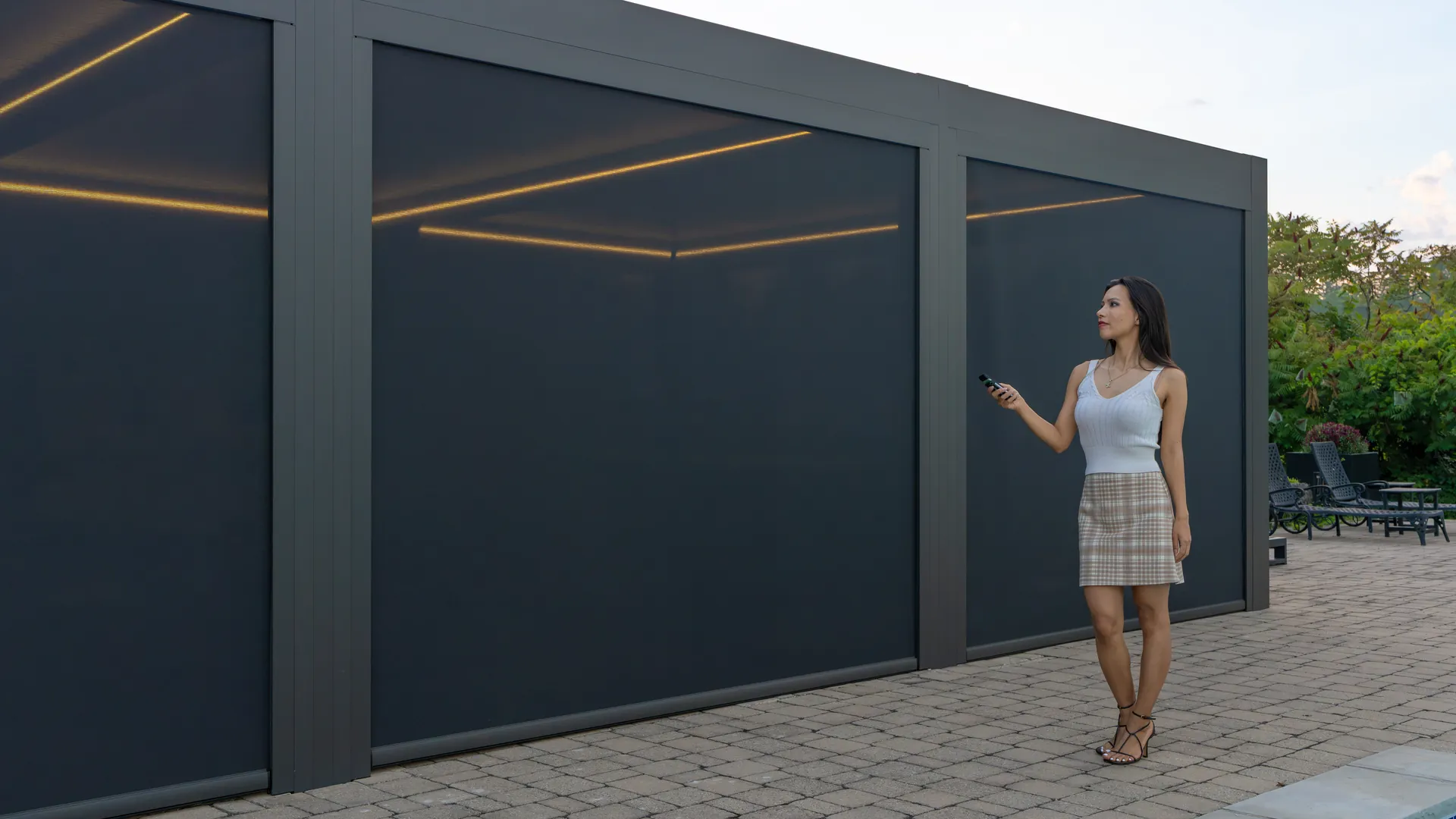 Woman using a handheld remote to control motorized ZIP screens on a dark grey aluminum pergola beside a pool