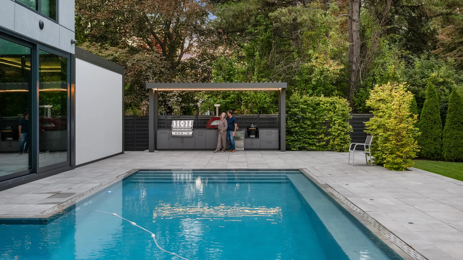 Backyard pool with a freestanding grey pergola over the outdoor kitchen at the far end of the patio