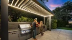 LED-lit louvers of an outdoor kitchen pergola at dusk, with a couple talking beside the stainless steel grill