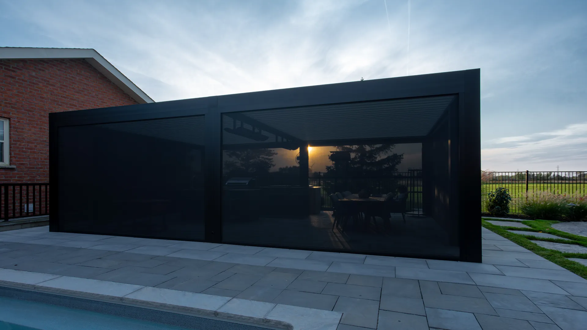 Low evening sun seen through outdoor ZIP screens on a black louvered pergola with a dining table beside a pool