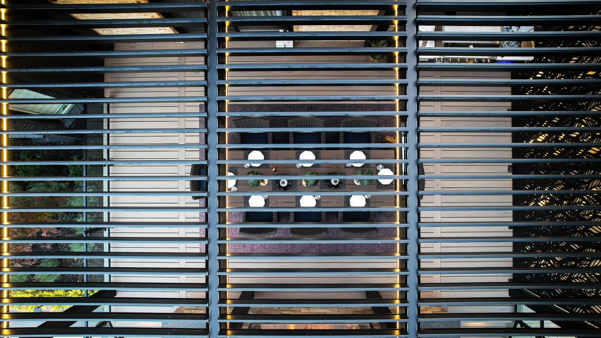 Top view through the open louvers of a pergola over a dining table set for dinner, with LED strips along the beams