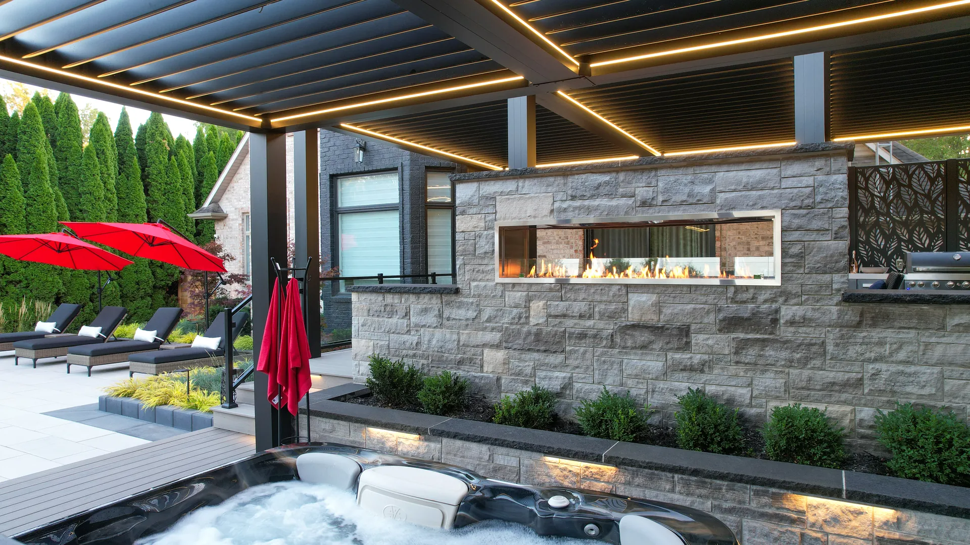 Black aluminum pergola with fireplace and grill seen from the hot tub, with the pool patio and red umbrellas on the left