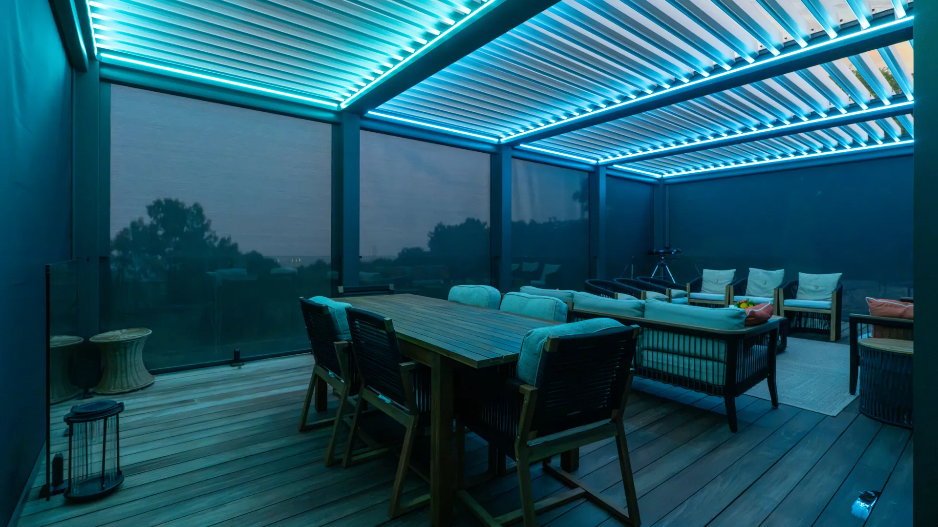 View from inside a louvered pergola through lowered ZIP screen fabric at dusk, with trees and the horizon still visible