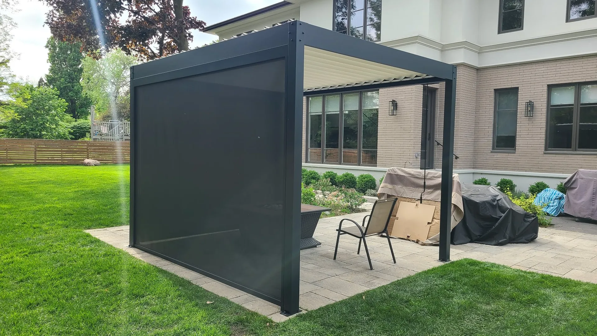 Dark grey louvered pergola with one ZIP screen lowered on a single side and a hand crank hanging from the roof beam