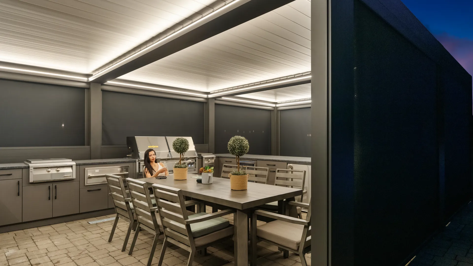 Louvered pergola over an outdoor kitchen and dining table at night, with ZIP screens lowered and warm LED lighting