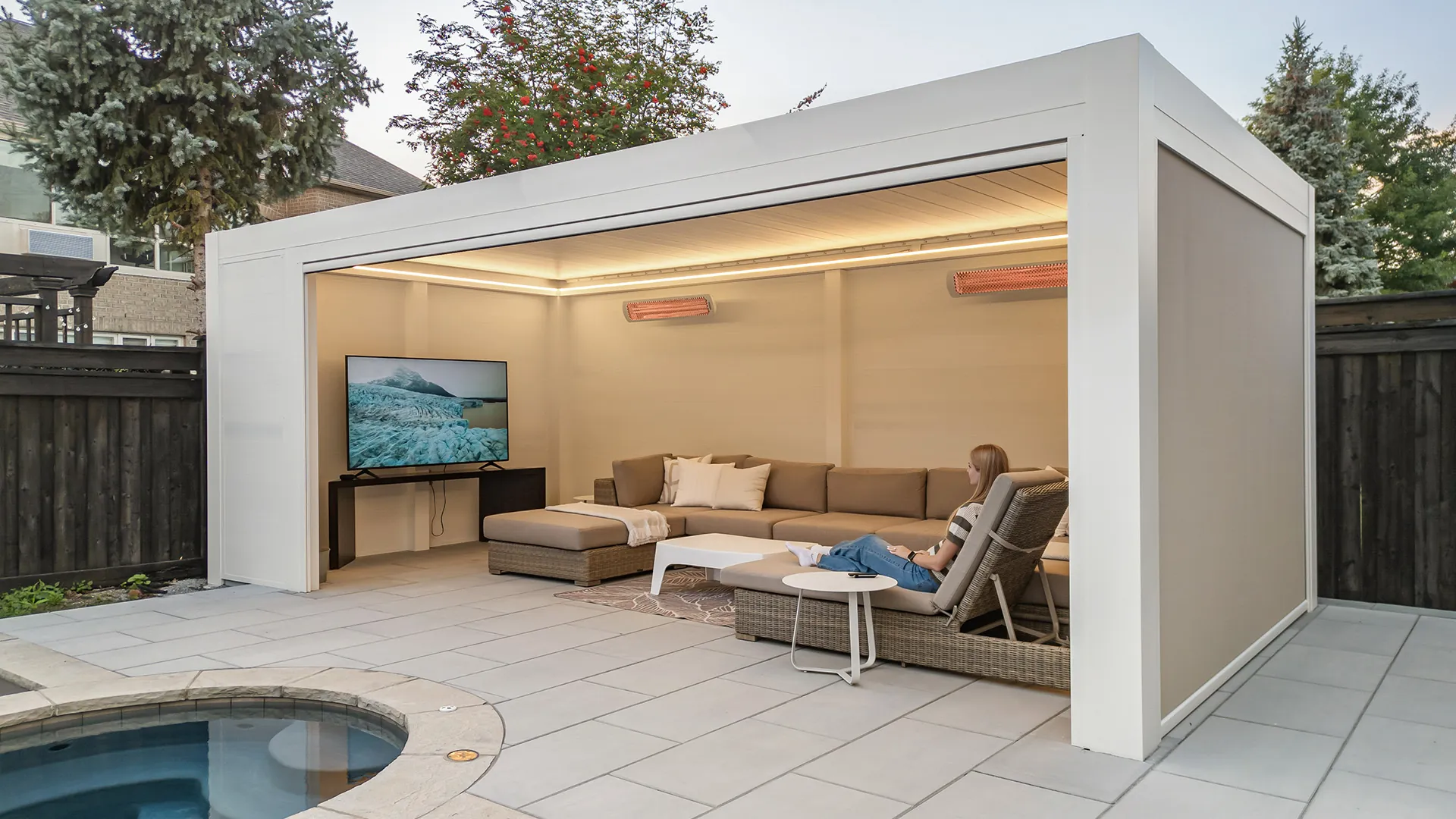White poolside pergola with warm LED lighting, two infrared heaters, a TV and a sectional sofa next to the pool spa