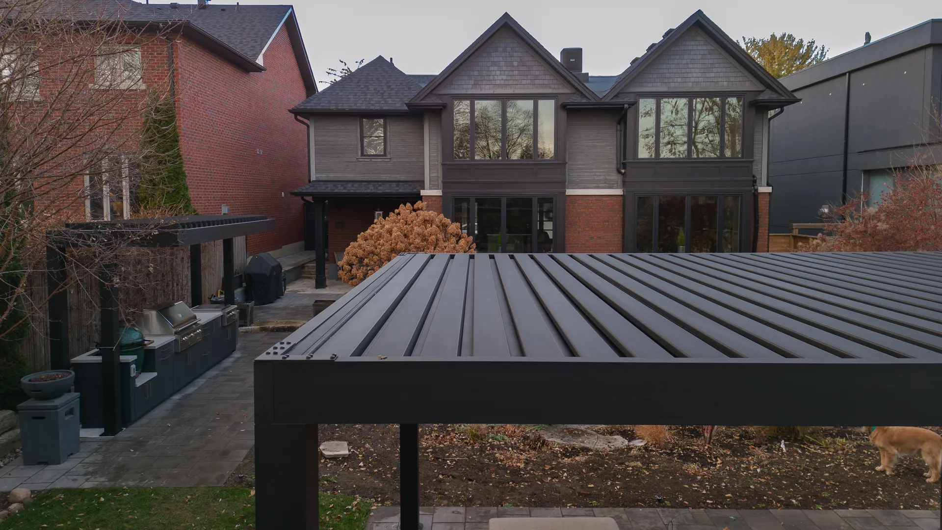 Two dark aluminum louvered pergolas in a backyard, one over an outdoor kitchen with a grill and one over the patio lounge