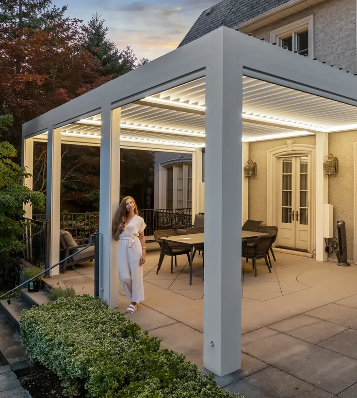 Woman under a white attached aluminum louvered pergola with LED lighting over a patio dining area beside the house